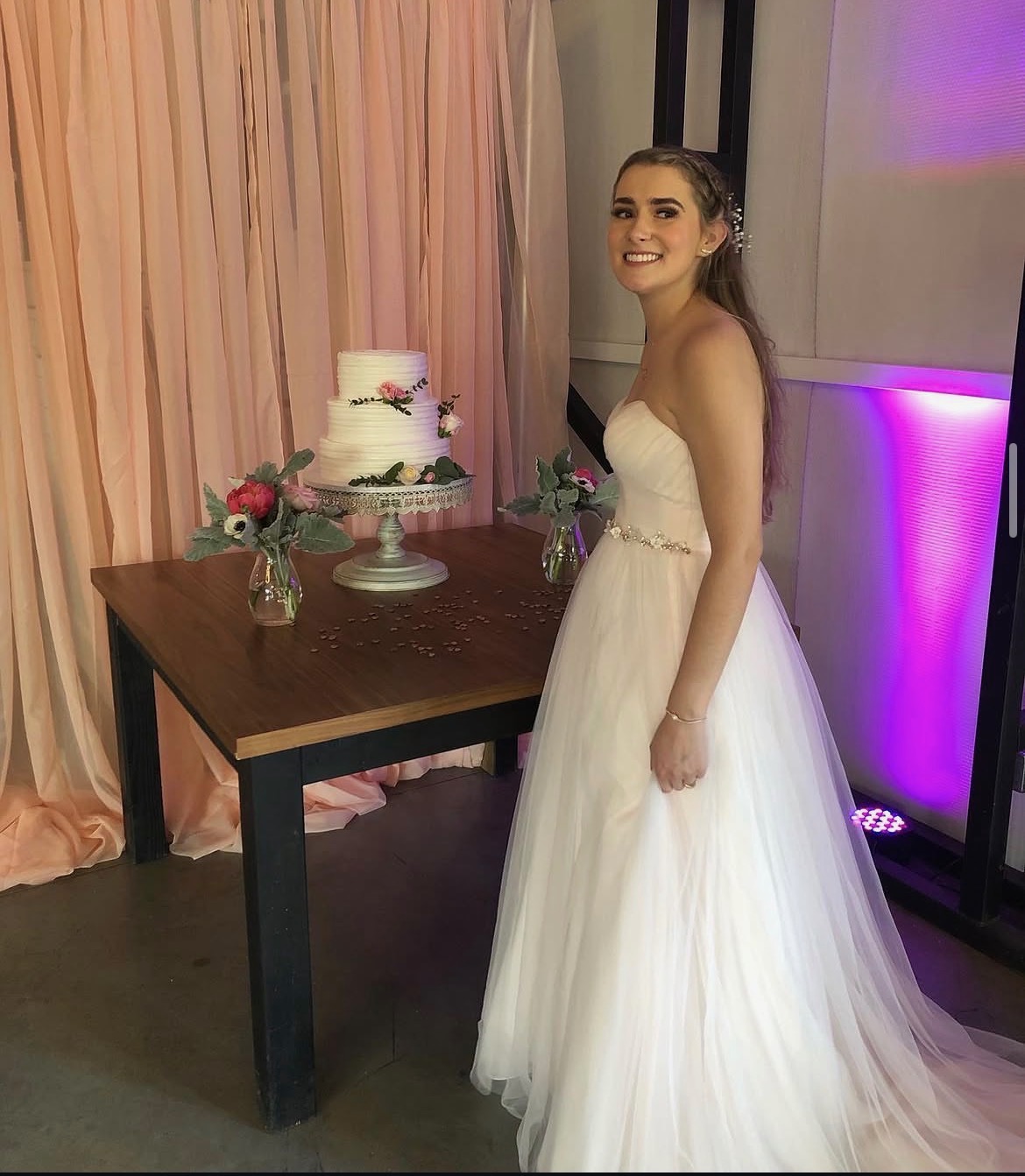 Bride posing next to wedding cake with NBT uplighting and pink draped backdrop